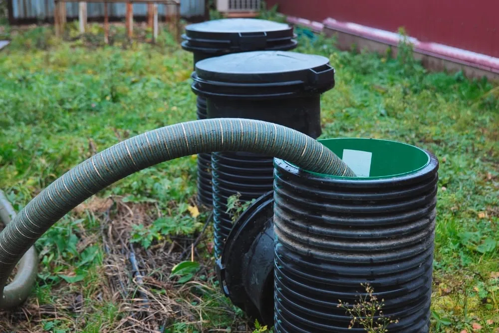 A large, flexible ribbed hose inserted into one of several black plastic septic system risers in a grassy backyard.